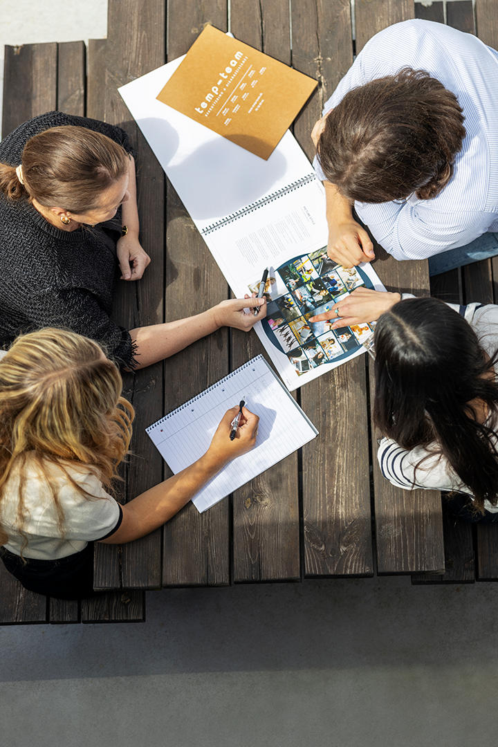 En mand og tre smilende damer sidder ved et bord-/bænkesæt udenfor og holder et møde med printet materiale på bordet.