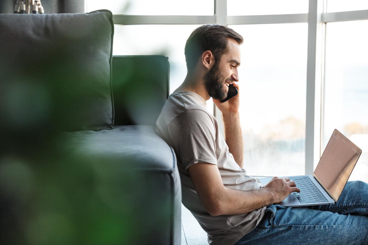 Leende ung man som pratar i telefon medan han arbetar på sin laptop hemma